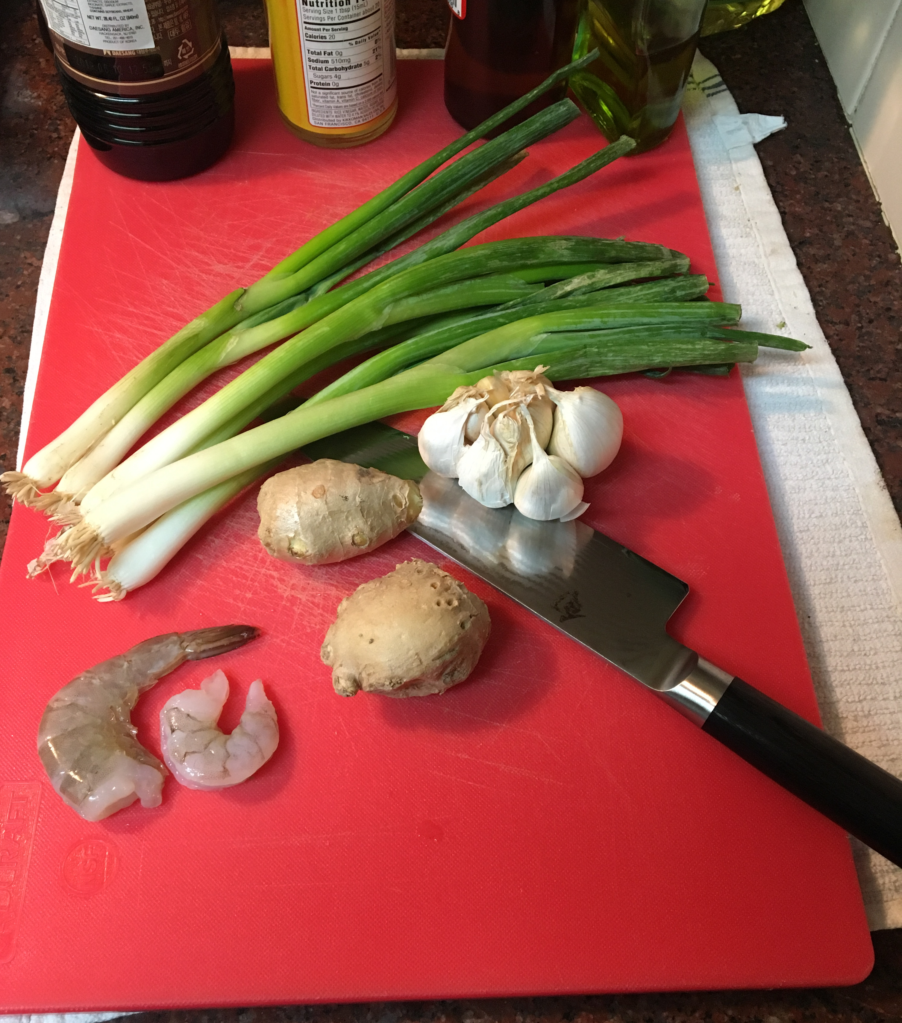 Everything you need! Fresh scallions, garlic, ginger and shrimp. Sesame oil, soy sauce, rice wine vinegar, olive oil. all on a cutting board with a chef knife. 